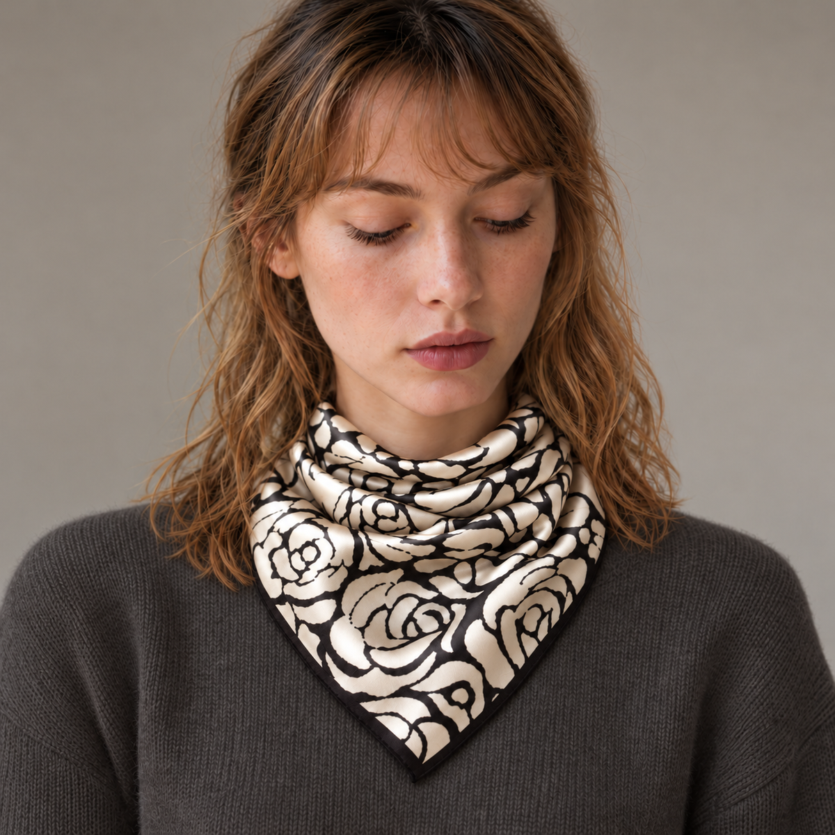 Le carré de soie Grandeur Roseraie, aux motifs noirs et écrus, noué au cou sur une maille gris anthracite.